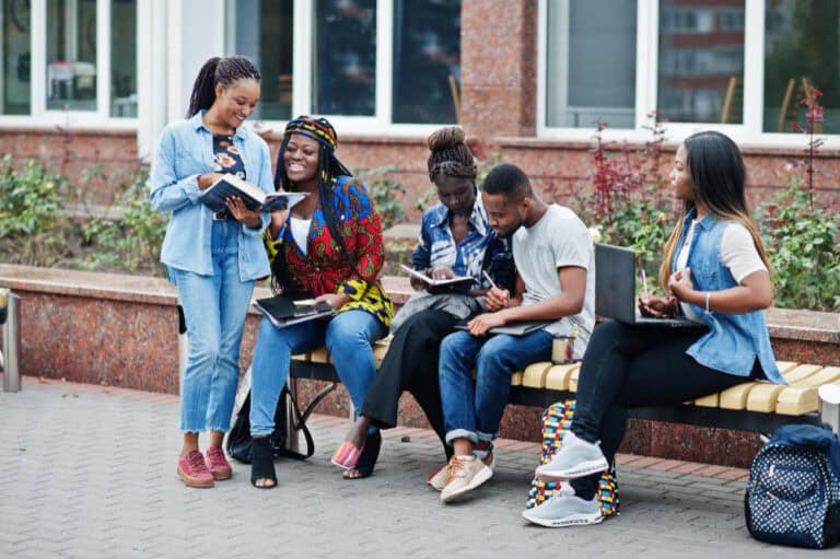 Group of diverse students sitting on a bench outside a brick building, reading and taking notes together.