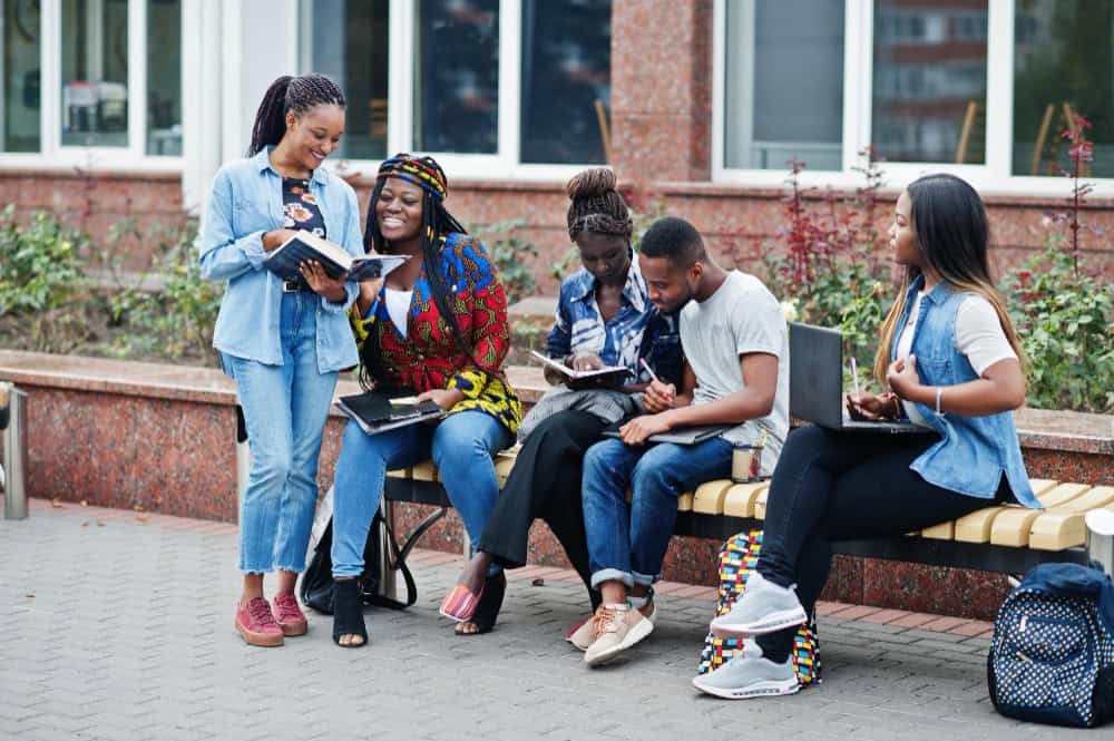 Group of diverse students sitting on a bench outside a brick building, reading and taking notes together.