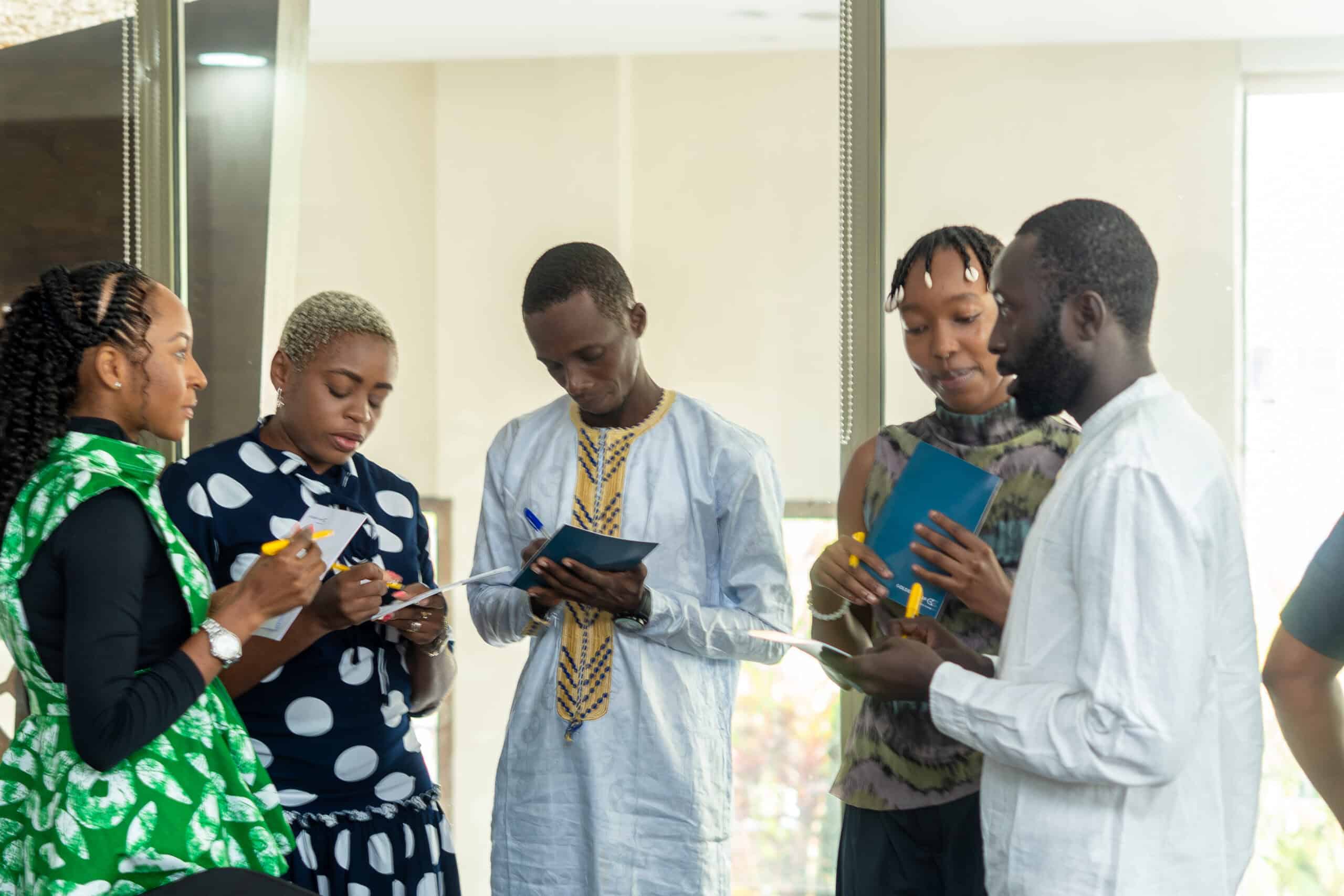 Group of five colleagues of diverse backgrounds standing by glass doors, reviewing papers and taking notes together.
