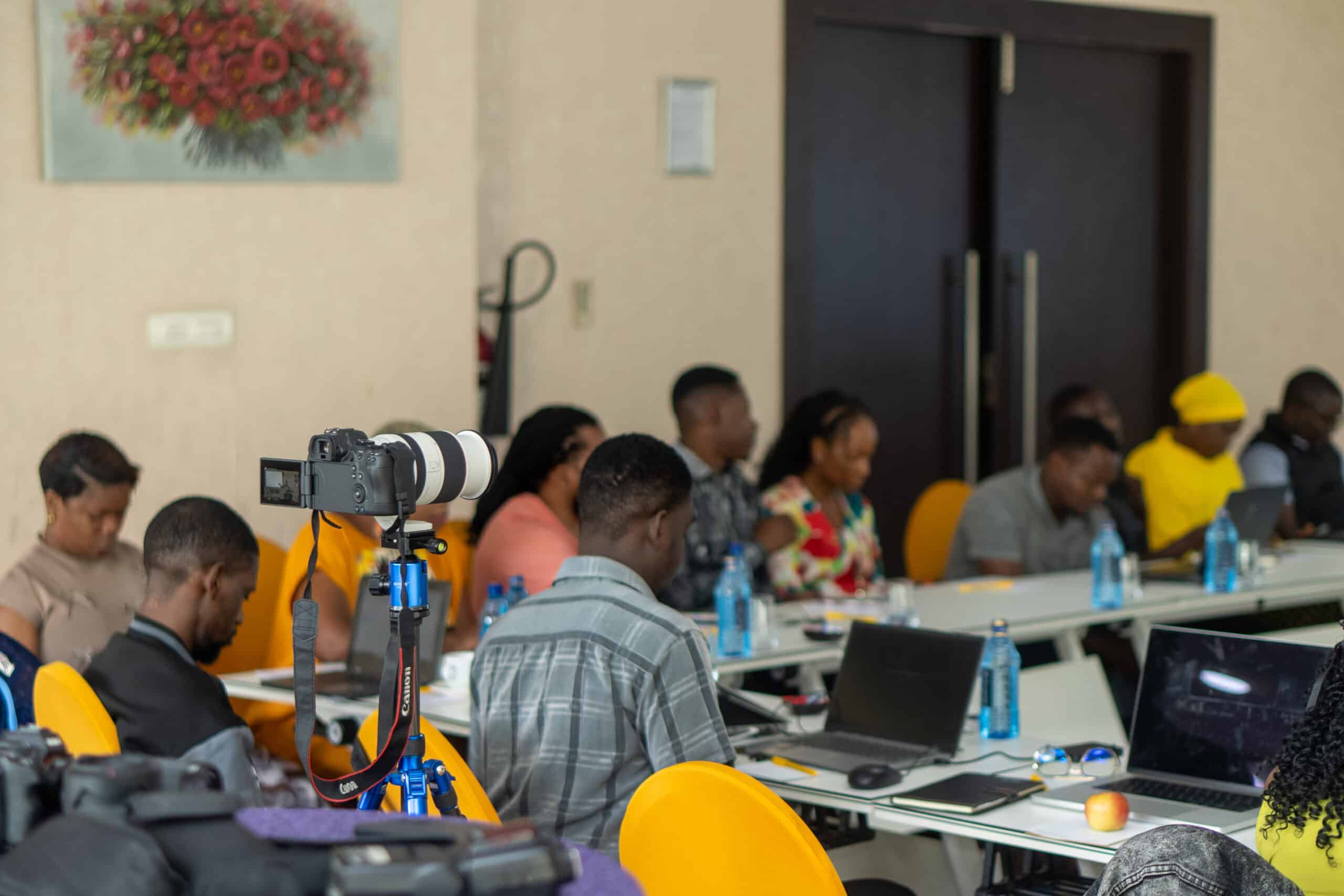 Conference room with attendees seated at a long table; a Canon camera on a tripod is in the foreground.