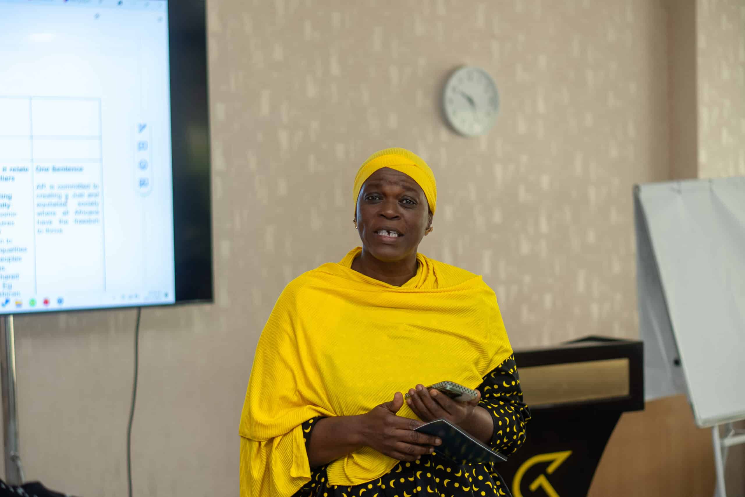 Woman in a bright yellow wrap speaking in a classroom, holding a smartphone, with a presentation screen and clock in the background.