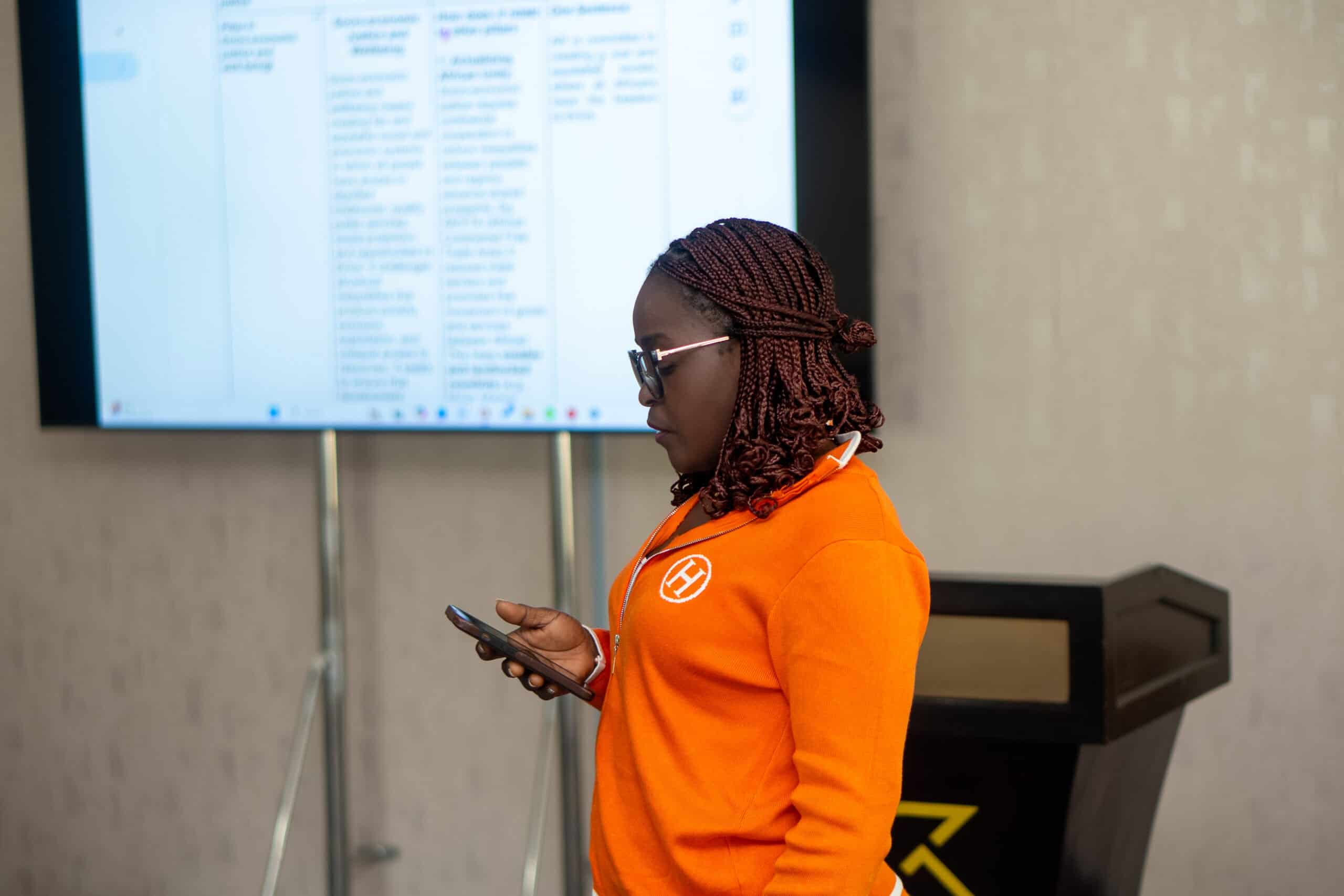 Side profile of a woman with braided hair in an orange shirt, holding a smartphone, standing near a large display with blurred text.