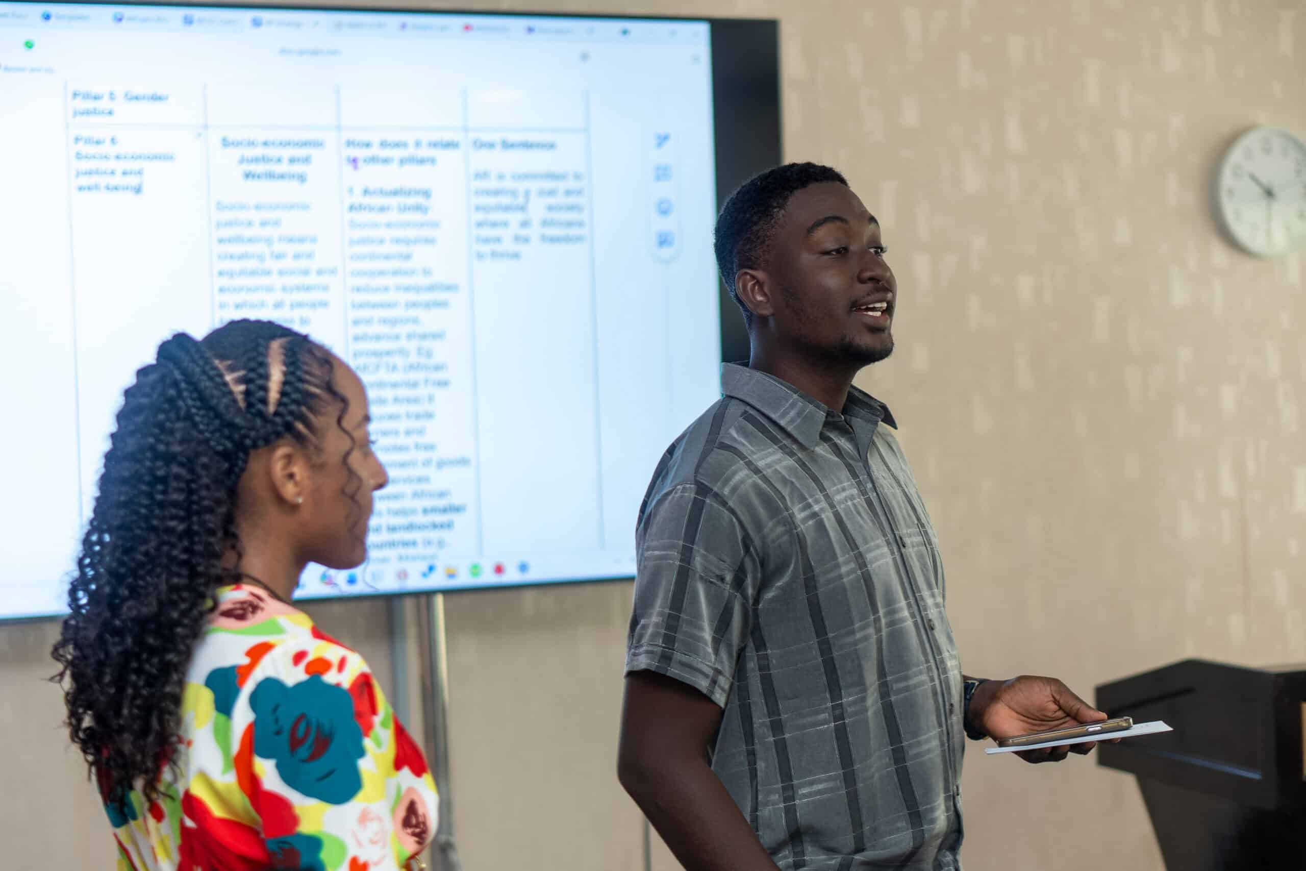 Two people presenting in a classroom: a man in a plaid shirt speaks while a woman with braided hair faces a large screen with a grid of text behind them.