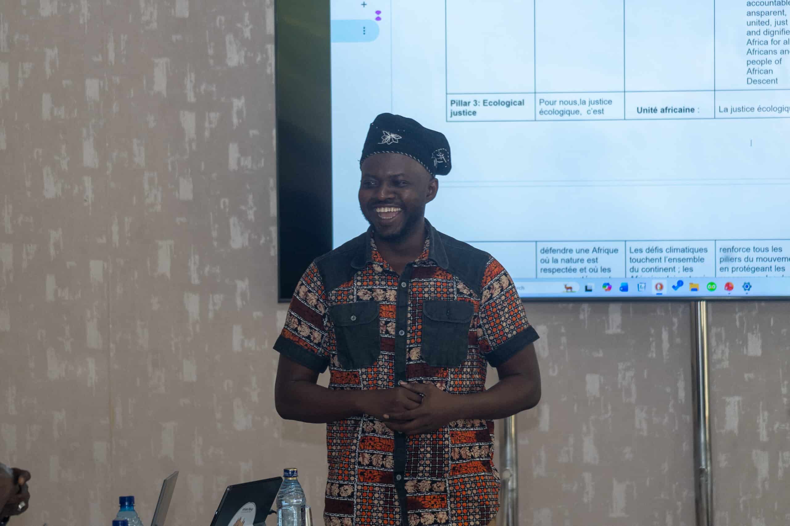 Smiling man in a patterned shirt and black headwrap presenting in front of a large screen with charts.