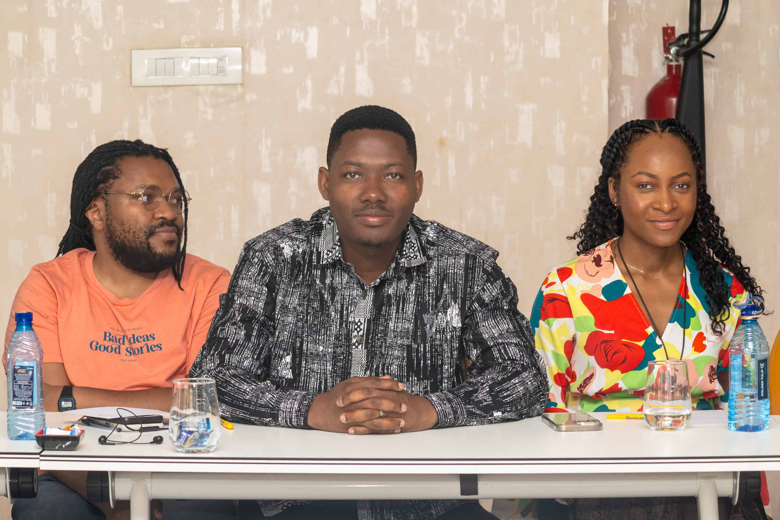 Three people sit at a table with water bottles and glasses, facing the camera; man in black patterned shirt in center flanked by a man in orange shirt and a woman in colorful dress.