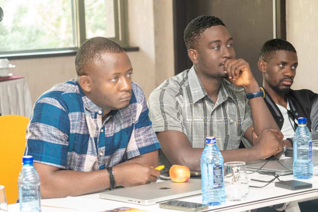 Three men sit at a table with water bottles, fruit, and laptops, engaged in a discussion at a meeting.