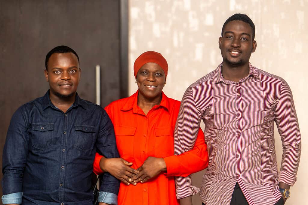 Three people posing together and smiling at the camera: a man in a denim shirt on the left, a woman in red with a headscarf in the middle, and a man in a striped shirt on the right.