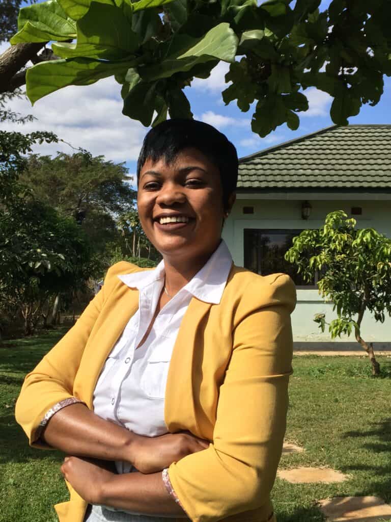 Woman smiling outdoors, wearing a yellow blazer over a white shirt, with trees and a house in the background.