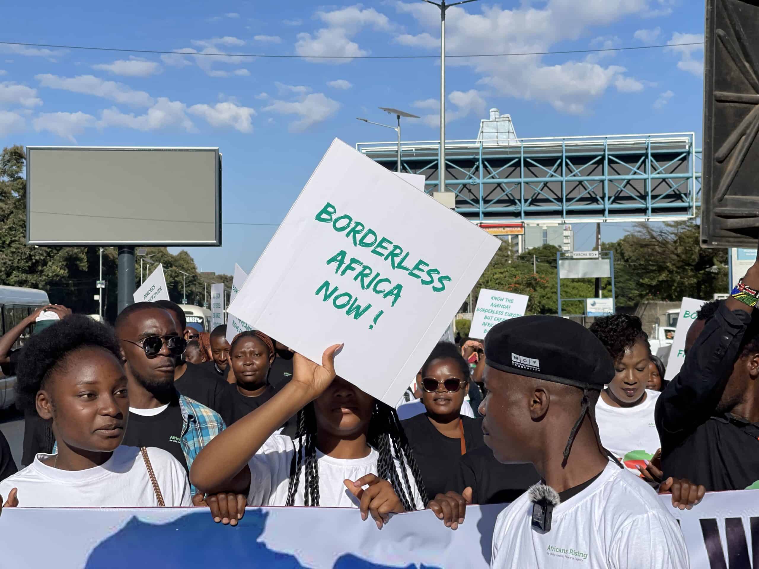 Crowd of protesters holding a sign reading 'Borderless Africa Now' during a street demonstration under a blue sky.