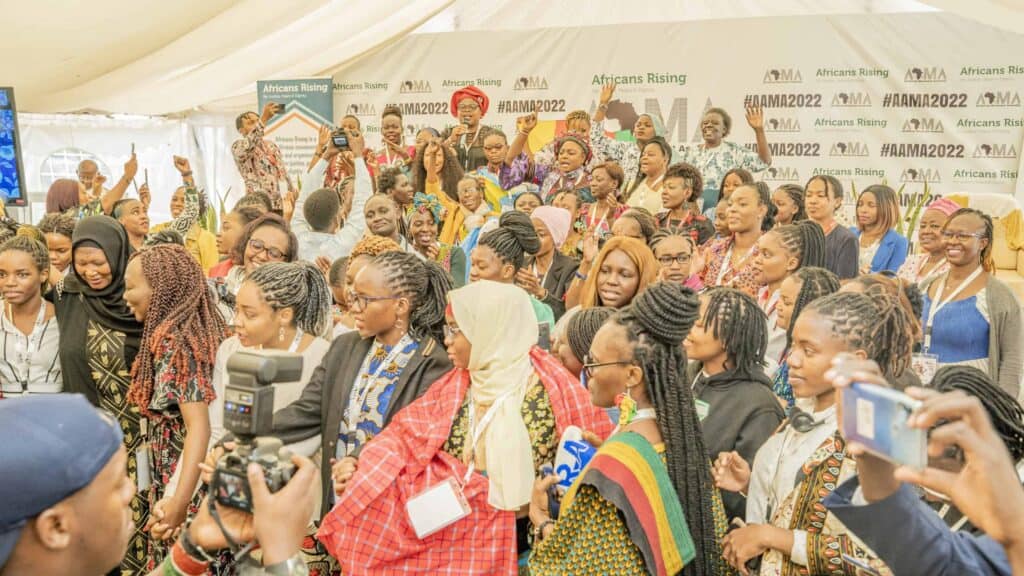 Large group of predominantly African women at a conference or rally, many raising hands and taking photos in front of a banner reading 'Africans Rising' and '#AAMA2022'.