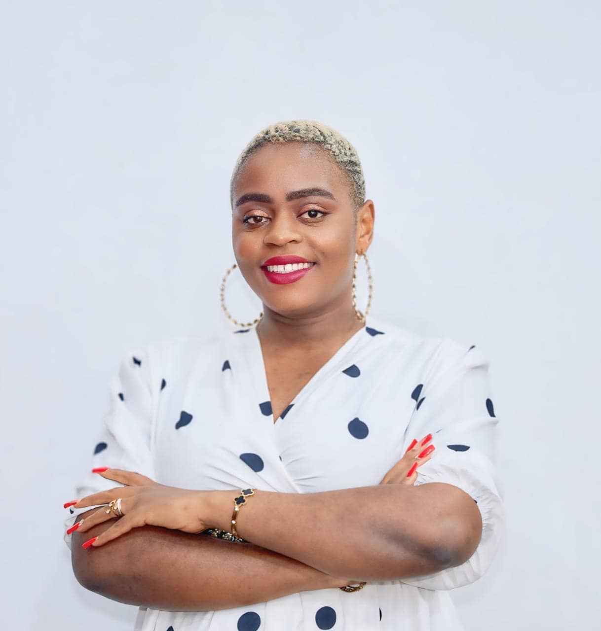 Portrait of a confident woman with short blonde hair, wearing a white polka-dot blouse, gold hoop earrings, red lipstick, and her arms crossed.