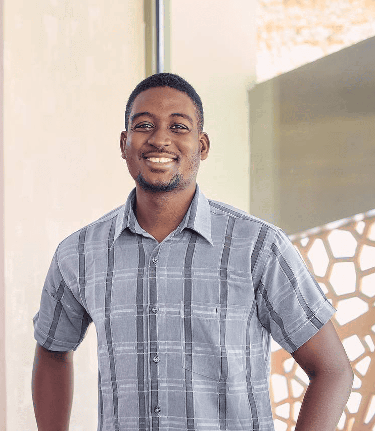 Smiling man in a gray plaid short-sleeve shirt posing indoors with a modern patterned wall in the background.