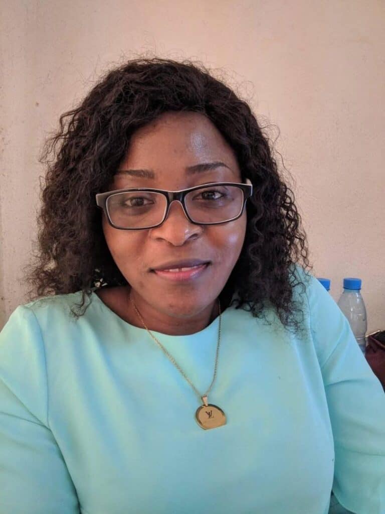 Woman with curly hair and glasses looking at the camera, wearing a light turquoise top.
