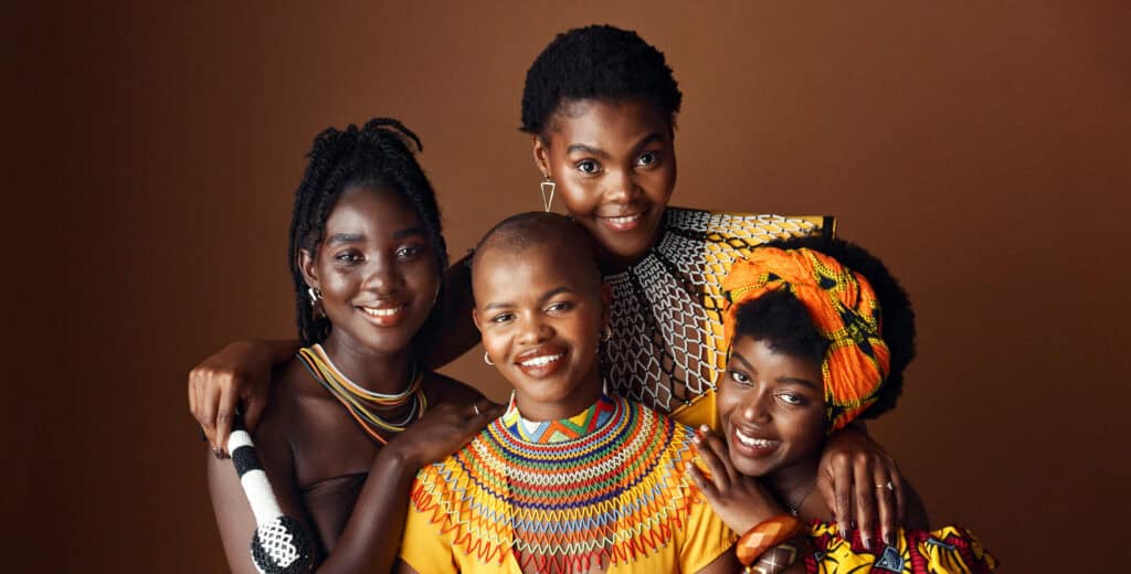 Four smiling women of African descent pose together, wearing colorful traditional outfits against a brown studio backdrop.