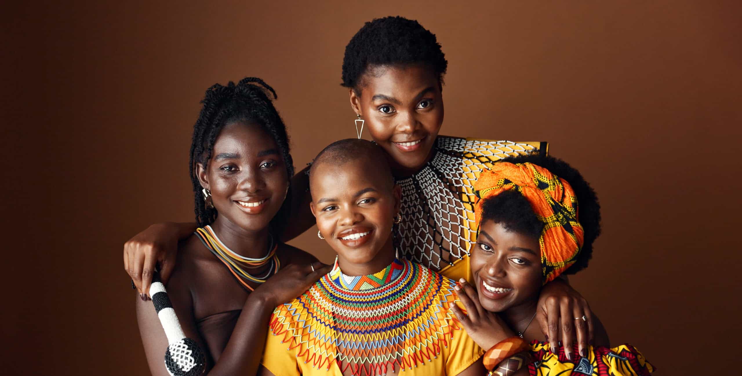 fashion-beauty-heritage-with-group-black-women-studio-brown-background-together-support-portrait-smile-culture-with-happy-african-people-clothes-tradition-community - Africans Rising - Africans Rising Four smiling women of African descent pose together, wearing colorful traditional outfits against a brown studio backdrop.