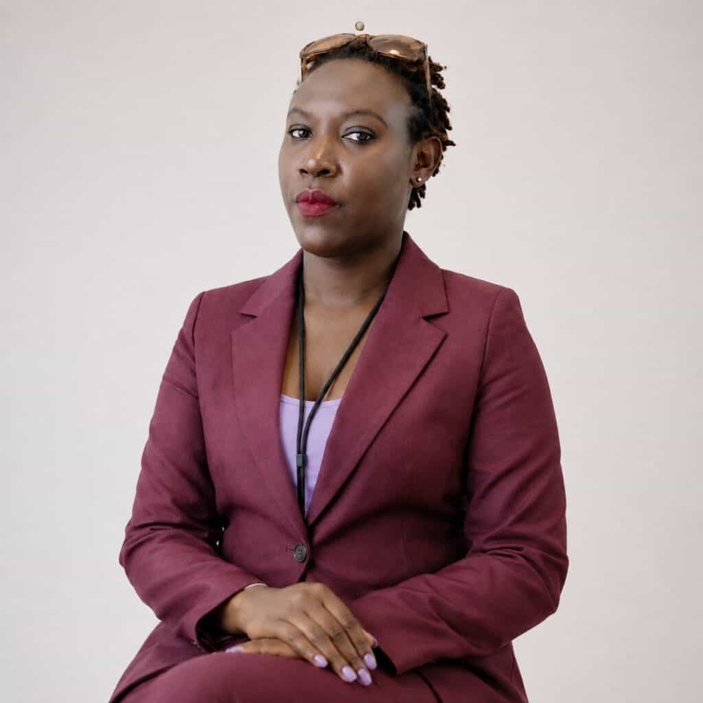 Portrait of a Black woman in a burgundy blazer and lavender top, seated with hands folded, sunglasses resting on her head.