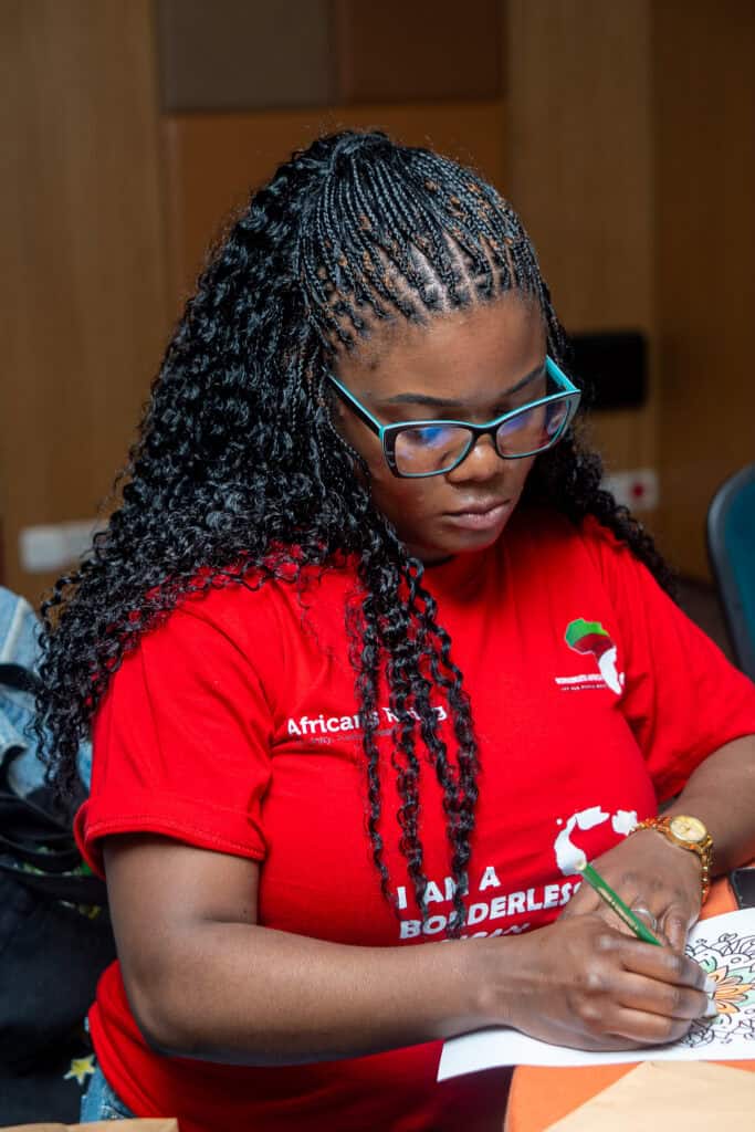 Woman with braided hair and teal-framed glasses coloring a page at a table.