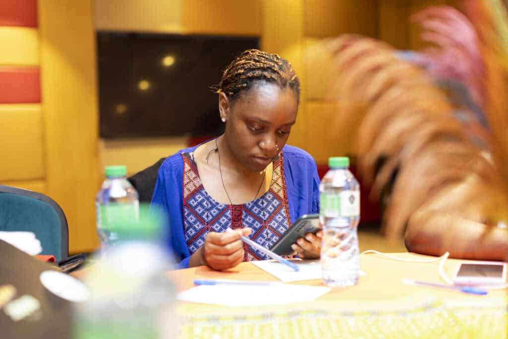 Woman with braided hair and a blue patterned top writes on papers at a table in a warm-lit room, looking down at her notes.
