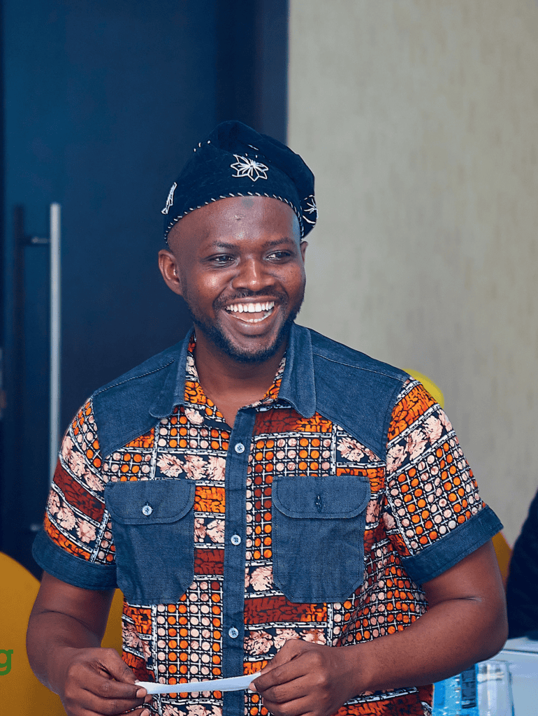 Smiling man wearing a dark head wrap and a colorful patterned shirt, holding a white strip of paper.