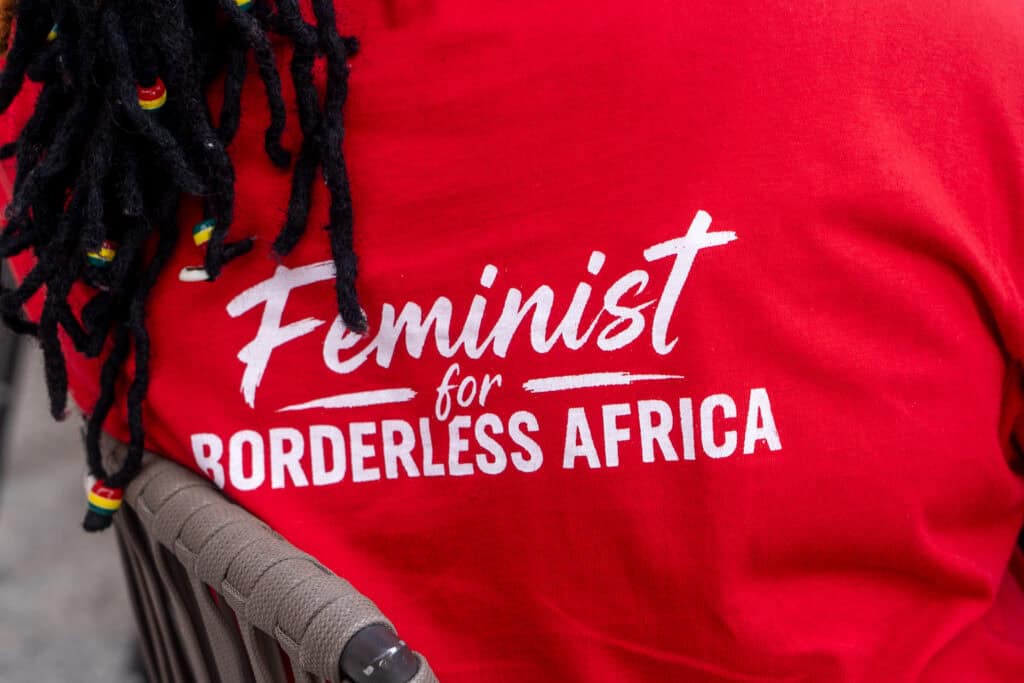 Close-up of a person wearing a red T-shirt with white 'Feminist for...' text and braided hair with colorful beads in the foreground.