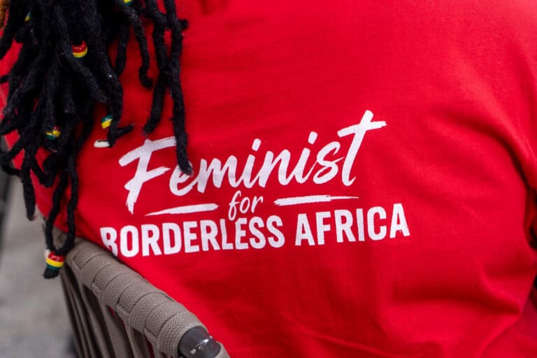 Close-up of a person wearing a red T-shirt with white 'Feminist for...' text and braided hair with colorful beads in the foreground.