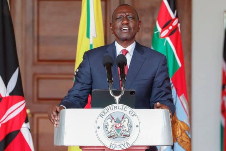 Male Kenyan official speaking at a podium with the Republic of Kenya seal, flags behind him.