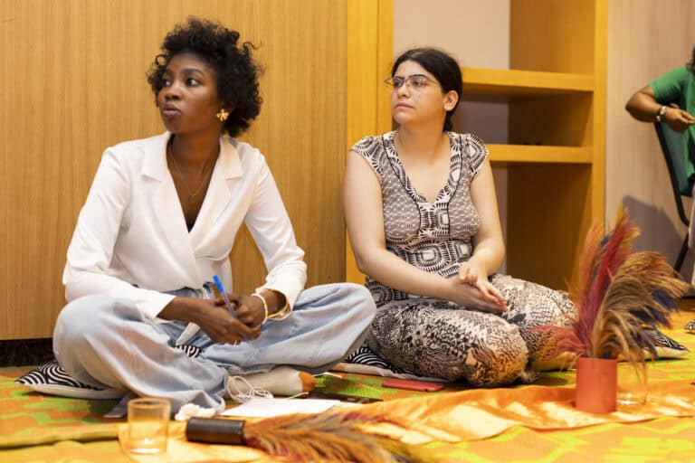 Two women sit on a colorful patterned blanket on the floor, leaning against a wooden wall with shelves; one wears a white blazer and jeans, the other a black-and-white patterned dress, both looking to the left as if listening.